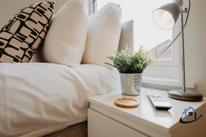 A neatly made bed with white bedding, a bedside lamp, a remote control, a small potted plant, and a wooden coaster on a white nightstand. - Flat B, sleeps 4, very central, 2nd Floor Derngate (West Northamptonshire)
