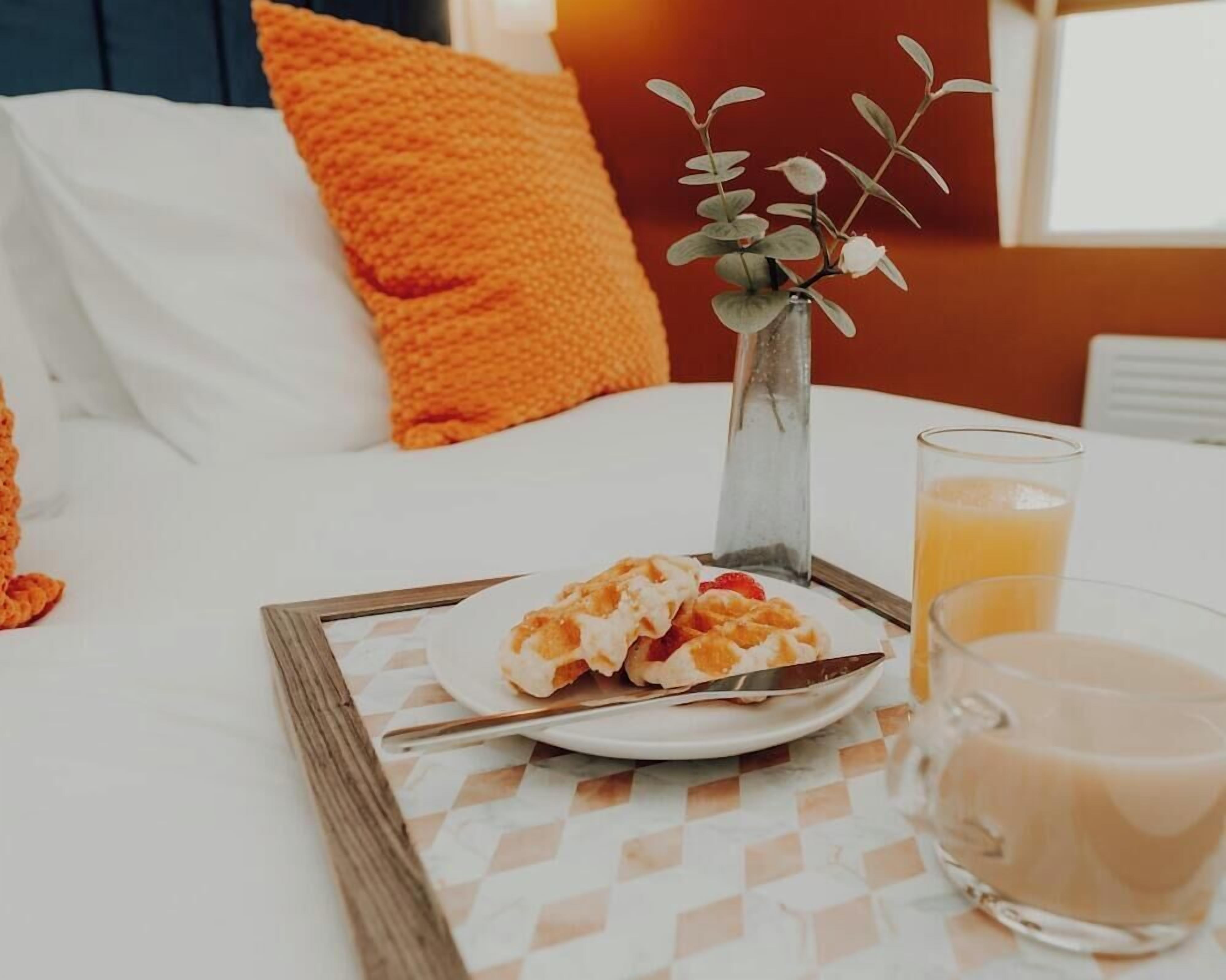 A breakfast setup on a bed with waffles, a glass of orange juice, and a vase with a plant.