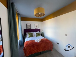 A hotel room with a bed, a nightstand, a lamp, and a wall with a logo. - Apt 4 Manor House - 1 Bed 1 bath sleeps 2 parking (Desborough)