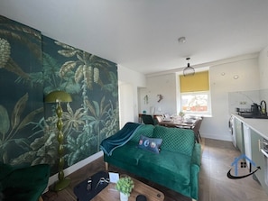 A compact living space with a green sofa, a dining table, a kitchen area, and a large window. - Apt 4 Manor House - 1 Bed 1 bath sleeps 2 parking (Desborough)