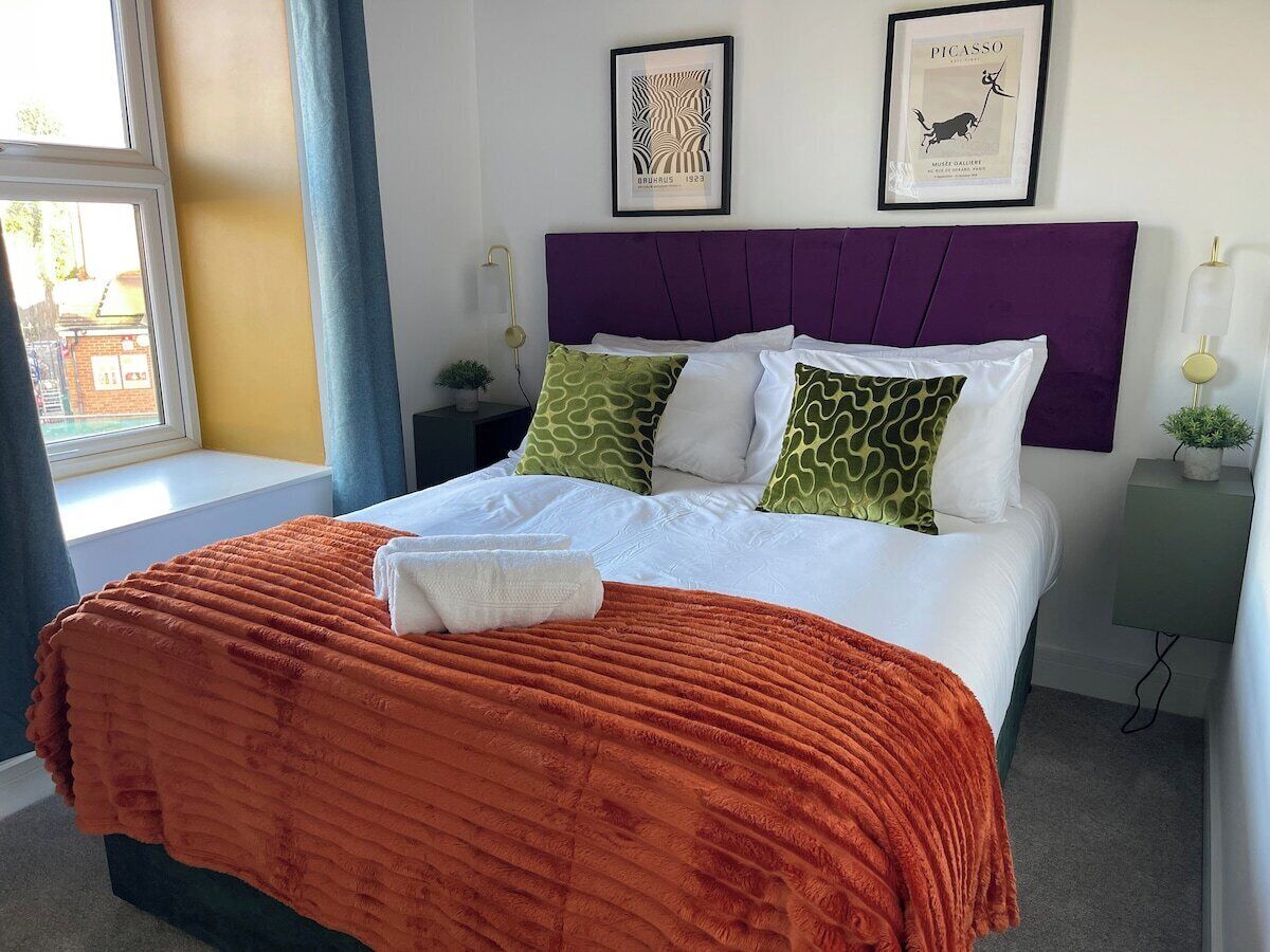 Ein ordentlich bezogenes Bett mit einem violetten Kopfteil, Kissen in Weiß und Grün gemustert und einer orangefarbenen Decke.