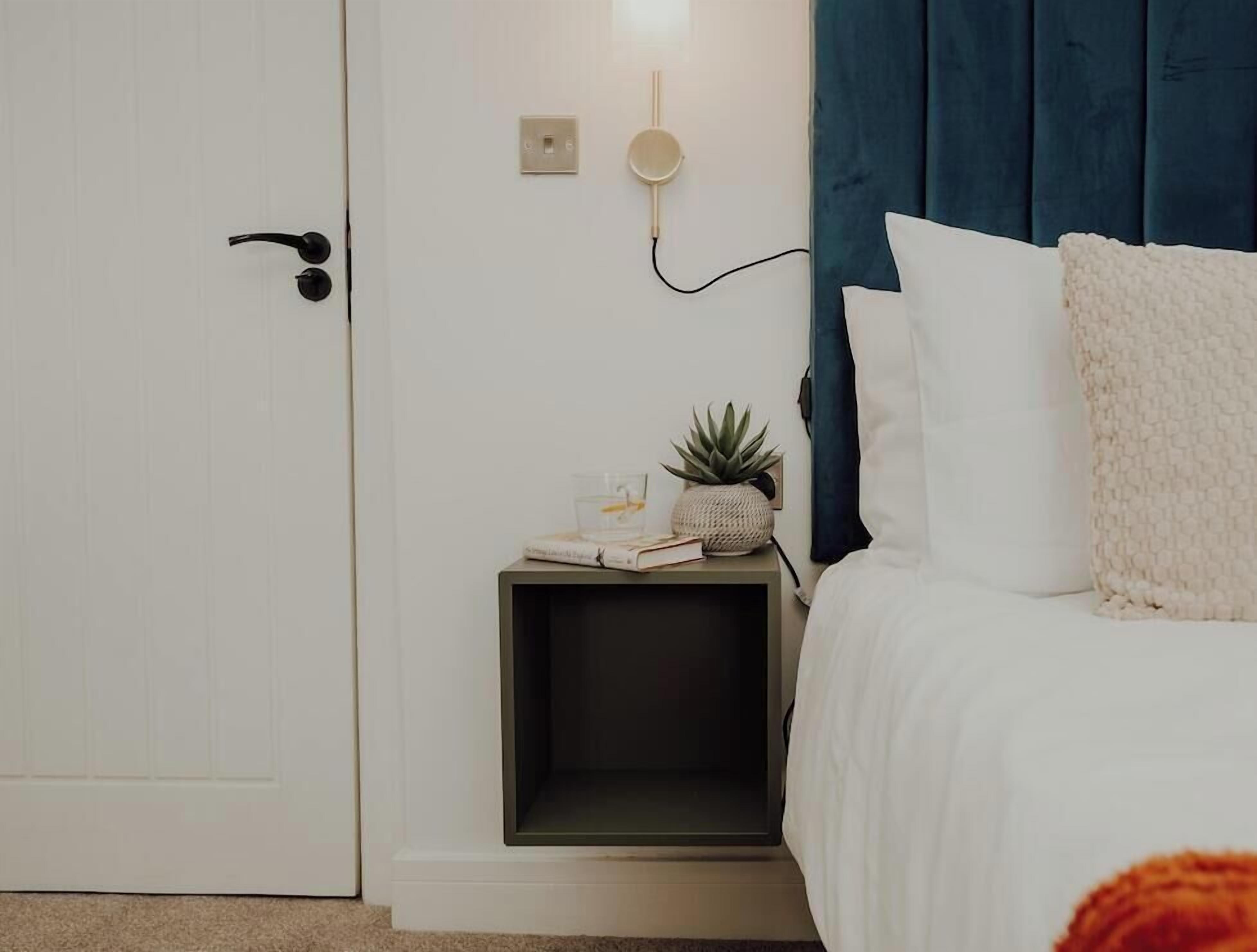 A bedroom with a bed, a bedside table, a nightstand, a wall-mounted lamp, and a door.