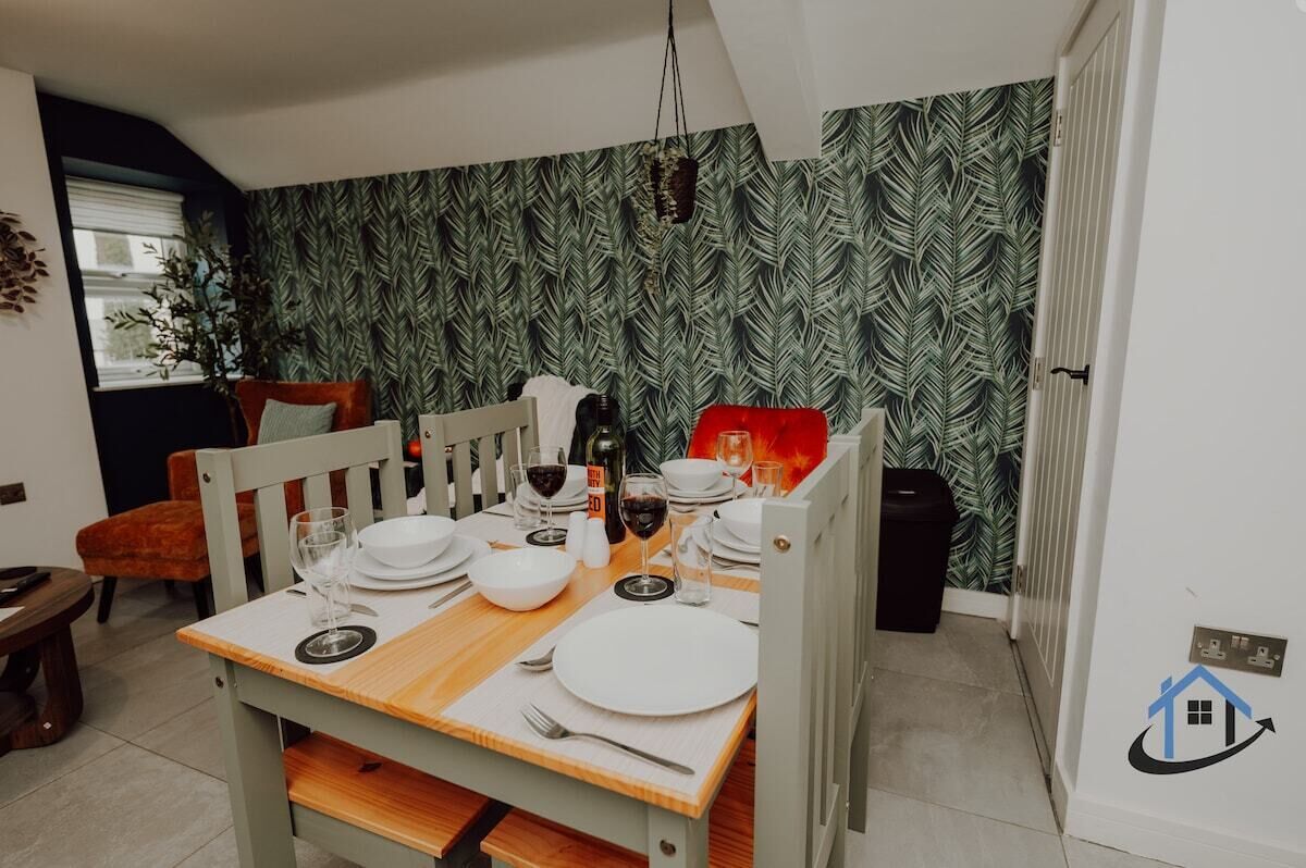 A dining area with a table set for four, a chair, a plant, and a patterned wallpaper.