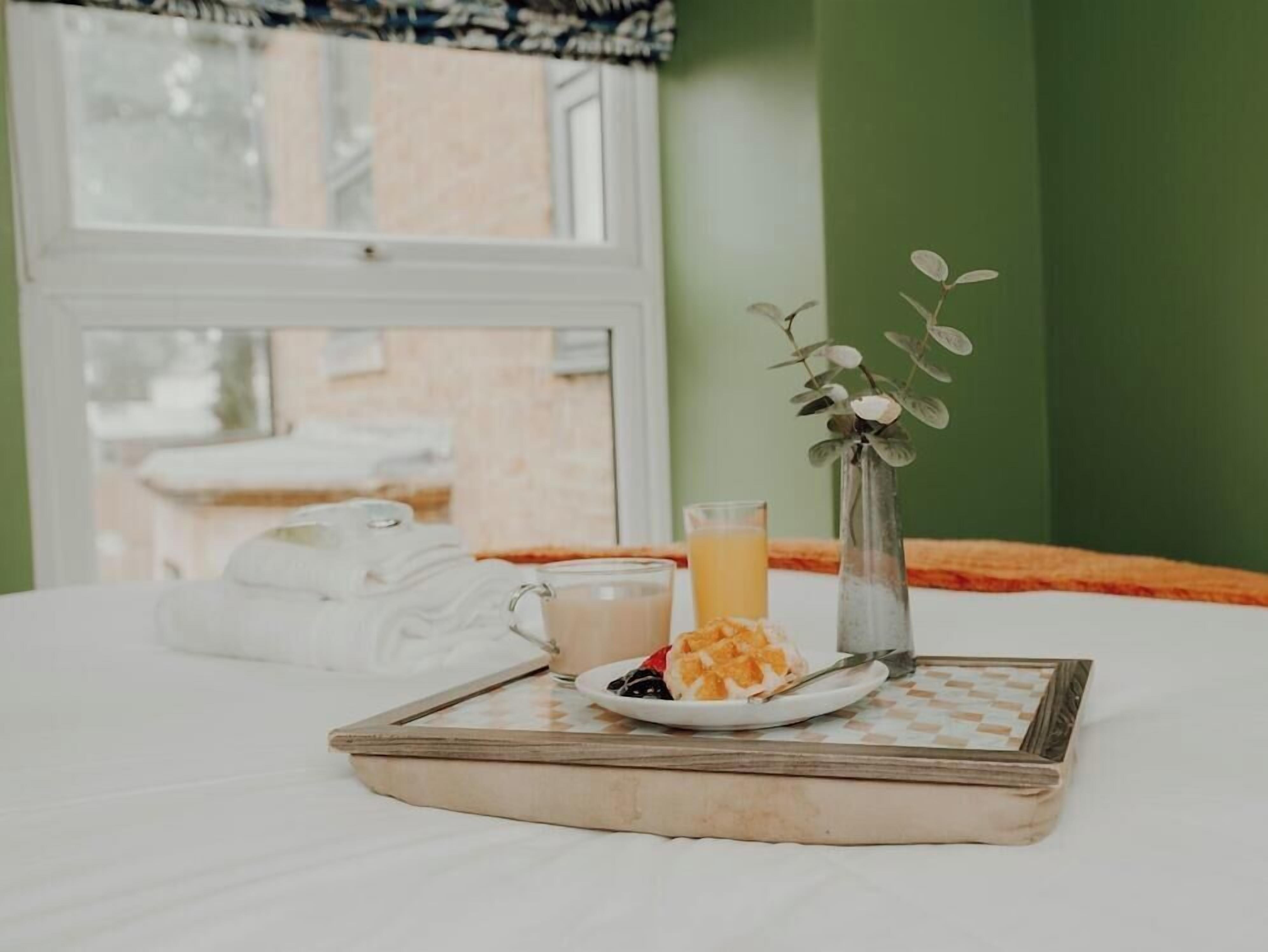 A breakfast tray with a glass of orange juice, a plate with a pastry and berries, and a vase with a plant on a bed with a window in the background.