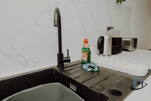 Een keuken met een zwarte spoelbak, een kraan, een Fairy afwasmiddel fles en een wit aanrecht.