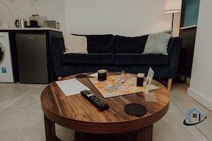Een moderne woonkamer met een donkerblauwe bank, een ronde houten salontafel en een keuken op de achtergrond.