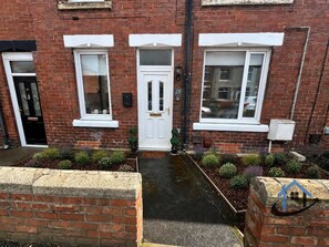 A brick house with three windows, a front door, and a small garden area. - Flat, sleeps 4, central, parking, Ground Floor (Bedlington)