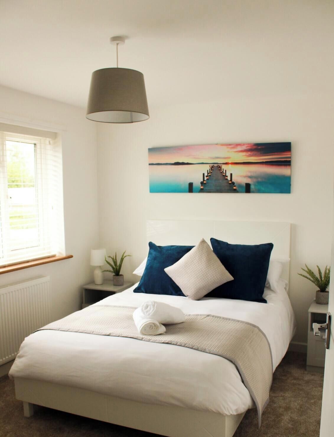 A bedroom with a bed, a nightstand, a lamp, a window, and a wall-mounted painting.