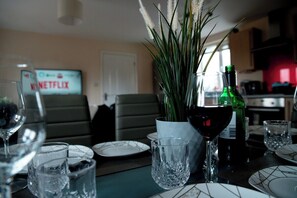 A dining table set with plates, glasses, and a bottle of wine, with a plant and a Netflix sign in the background.