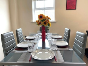 A dining table set for six with a vase of sunflowers, grey upholstered chairs, and a window in the background.