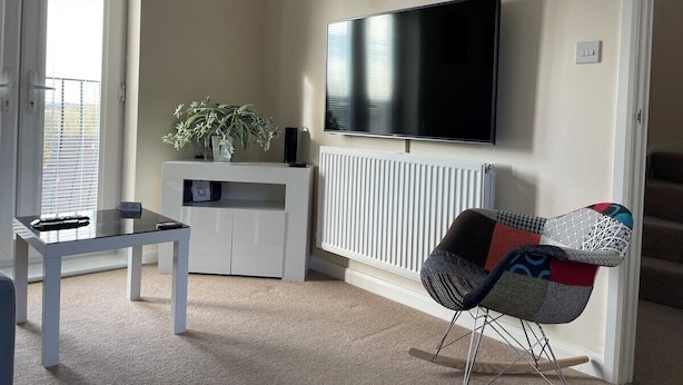 Una sala de estar con un televisor de pantalla plana montado en la pared, un mueble blanco debajo, una mesita con un control remoto, un sillón colorido y una planta en maceta.