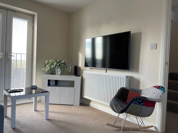 A living room with a flat-screen TV mounted on the wall, a white cabinet below it, a small table with a remote control, a colorful chair, and a potted plant.