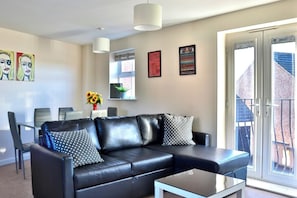 A modern living room with a black leather sofa, a glass coffee table, and a dining area with chairs and a table. There are framed artworks on the walls and a large window with a view of the outside.