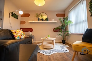 A modern living room with a sofa, a round coffee table, a potted plant, and a window with blinds. - Minchin House 3 bed parking sleeps 8 (Northamptonshire)