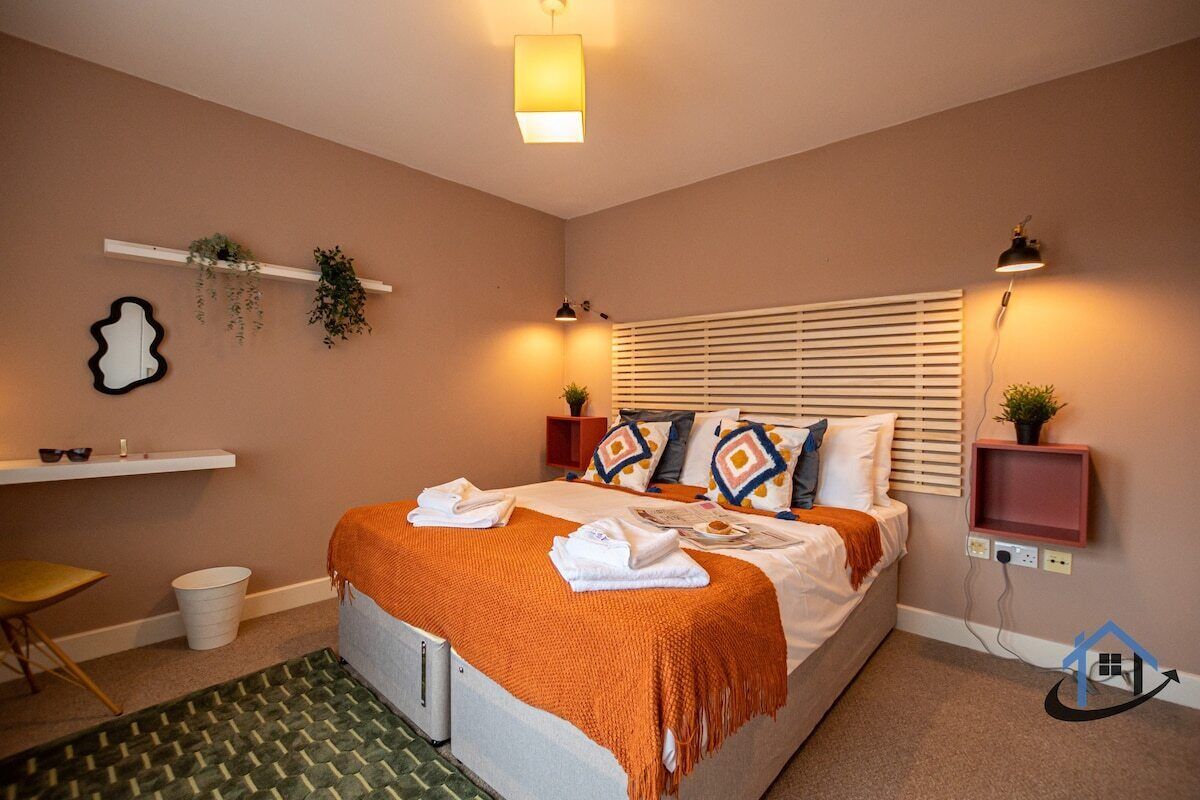 A bedroom with a bed, bedside tables, a hanging plant, a shelf, and a desk.