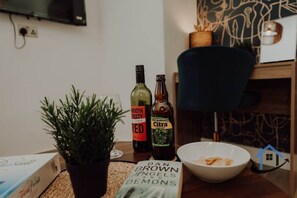 A table with a potted plant, a book titled 'Dan Brown Angels and Demons,' a bowl of snacks, and two bottles of wine. - Apt 6 Manor House 1 Bed 1 bath sleeps 2 parking (Desborough)