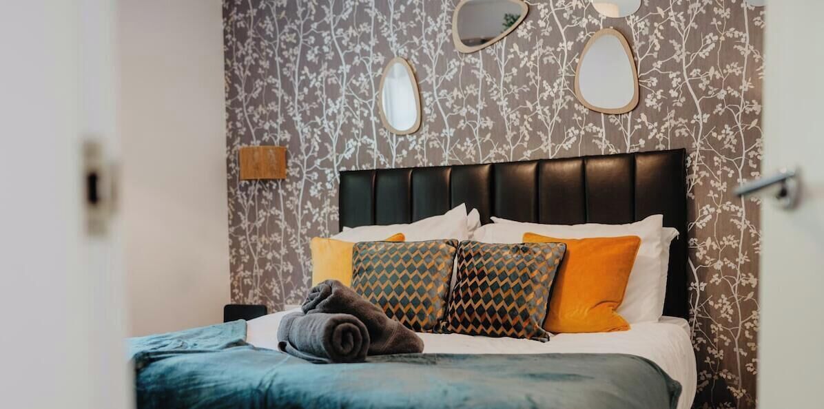 Un lit bien fait, avec une tête de lit à motifs, un miroir fixé au mur et une serviette posée sur le lit.