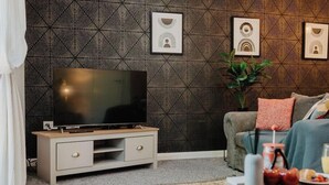 Ett modernt vardagsrum med en platt-TV på ett träställ, en soffa med kuddar, ett soffbord med ett bricka fullt med mat och en vägg med ett mönster.