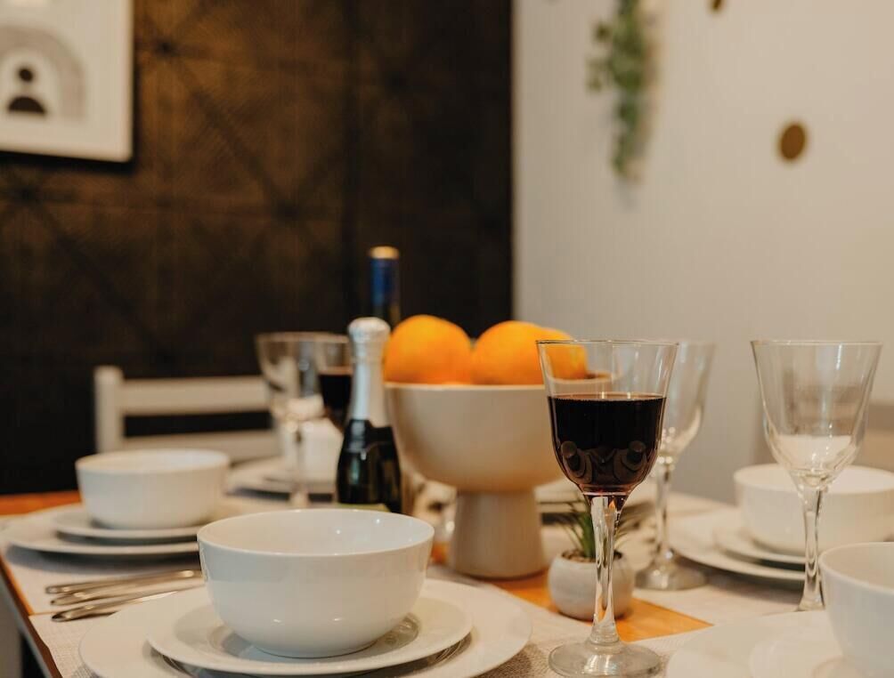 A dining table set with white plates, bowls, and wine glasses, with a bowl of oranges and a bottle of wine in the background.