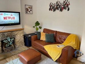 A living room with a leather sofa, a television mounted on the wall, a fireplace, and a decorative wall piece. - Petham Cottage 2 bed sleeps 6 (Stanwick)
