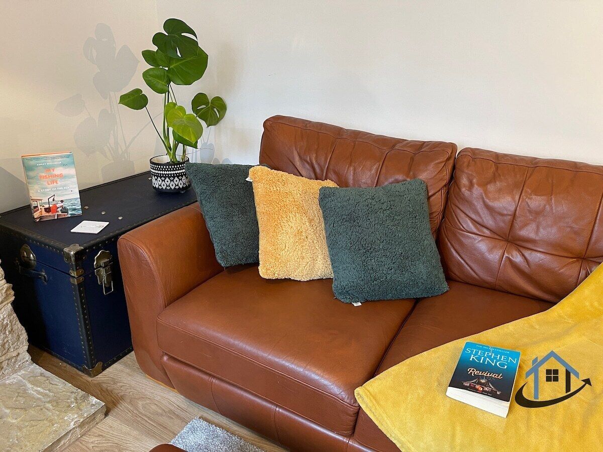 A brown leather sofa with two cushions, a small table with a book and a plant, and a suitcase.