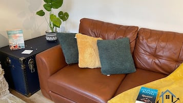 A brown leather sofa with two cushions, a small table with a book and a plant, and a suitcase.