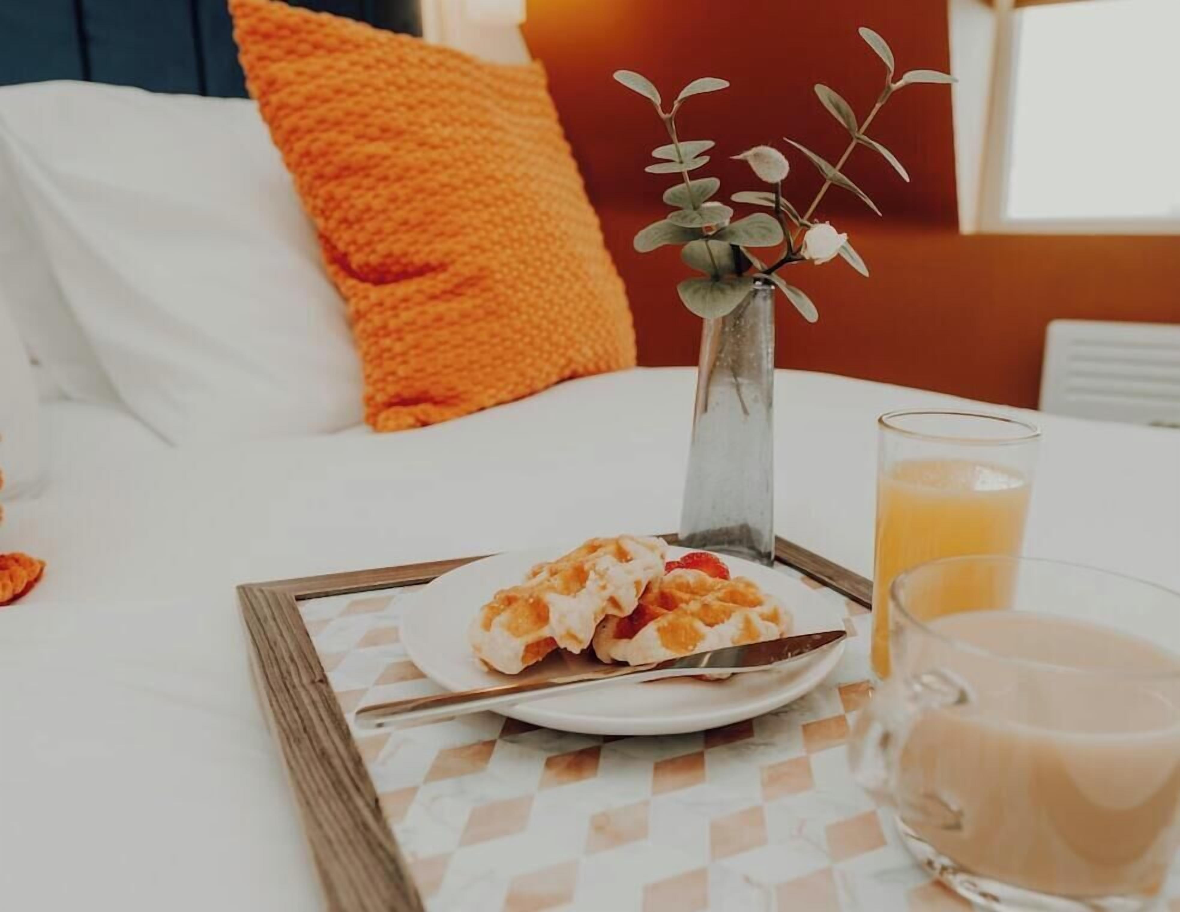 A breakfast tray with waffles, a glass of orange juice, and a glass of coffee on a bed with white bedding and an orange pillow.