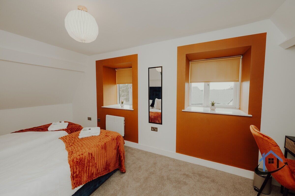 Quarto de hotel com cama, duas janelas com persianas, radiador, espelho fixado na parede e luminária suspensa.