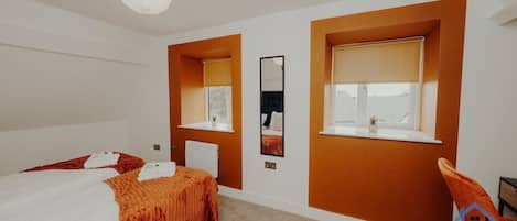 Una habitación de hotel con cama, dos ventanas con persianas, un radiador, un espejo fijado a la pared y una lámpara colgante.