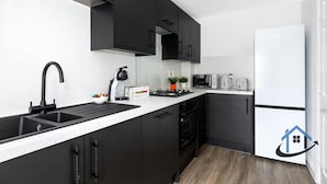 A modern kitchen with black cabinets, a white countertop, a sink, a stove, a microwave, a toaster, and a refrigerator. - Bluebell House, sleeps 7, parking x2, garden (Corby)