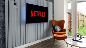 A modern living room with a mounted television displaying 'NETFLIX' in red letters, a comfortable orange armchair, and a glass coffee table. - Bluebell House, sleeps 7, parking x2, garden (Corby)