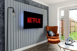 Ett modernt vardagsrum med en väggmonterad tv som visar ordet ”NETFLIX” med röda bokstäver, en bekväm orange fåtölj och ett soffbord i glas.