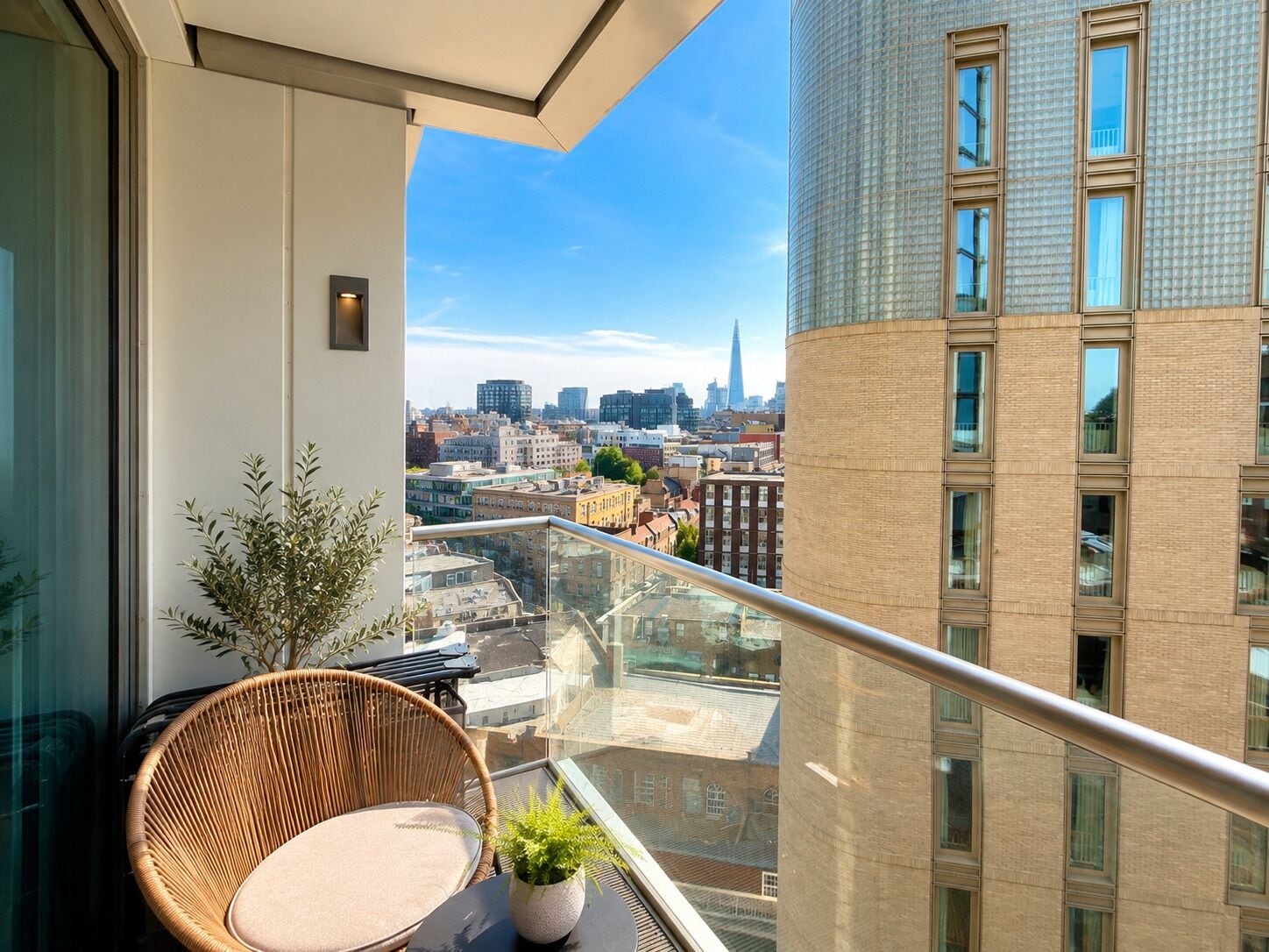 Ein Balkon mit einem Korbstuhl und einem Blumentopf, mit Blick auf eine Stadtlandschaft mit Gebäuden und blauem Himmel.