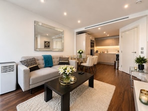 A modern living room with a sofa, coffee table, and a dining area with chairs. There is a kitchen in the background. - Luxury City View AC Balcony Rooftop Panoramic - Fast Wi-fi, near Sky Garden (London)