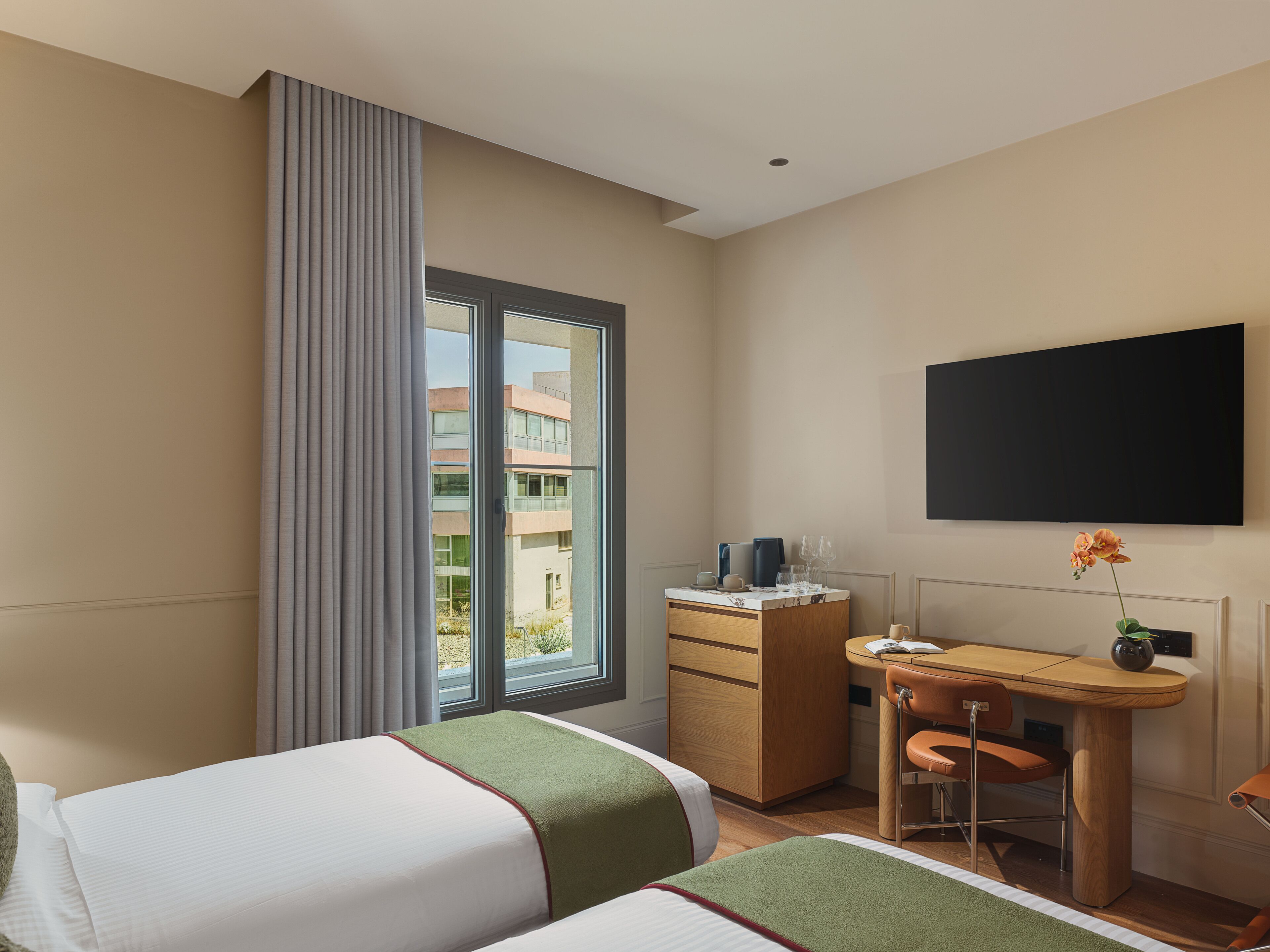 Quarto de hotel com duas camas, uma escrivaninha com cadeira, televisão de tela plana e janela com cortinas.