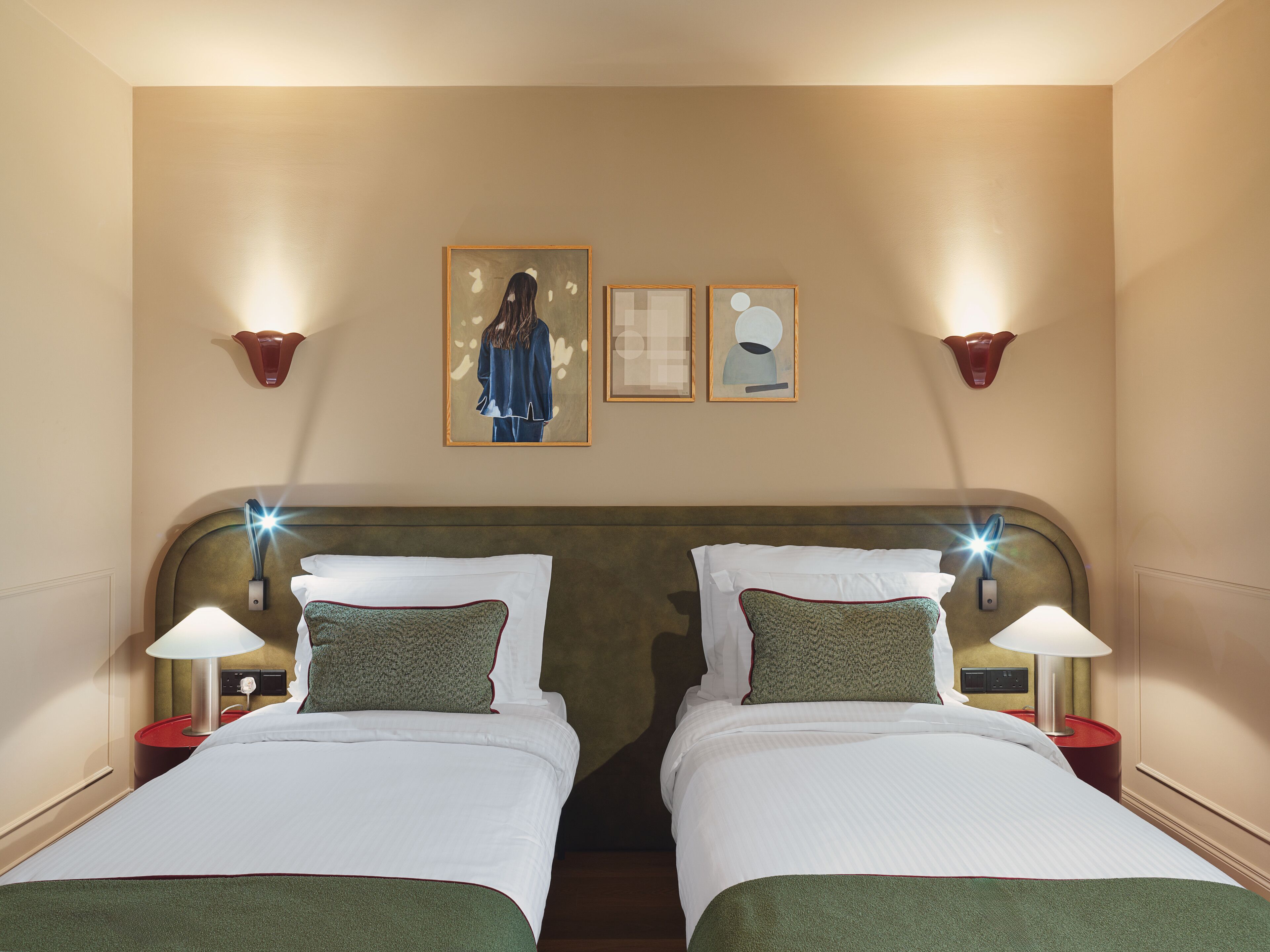 Ein Hotelzimmer mit zwei Betten, jeweils mit einem Kopfkissen und einer Lampe. An den Wänden befinden sich Leuchten und über den Betten Kunstwerke in Bilderrahmen.