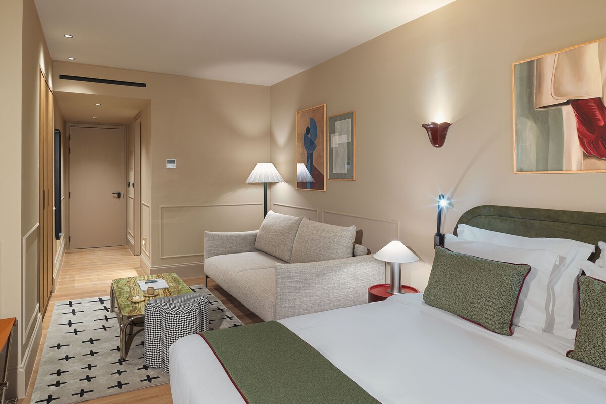 A hotel room with a bed, a sofa, a small table, a lamp, and artwork on the walls.
