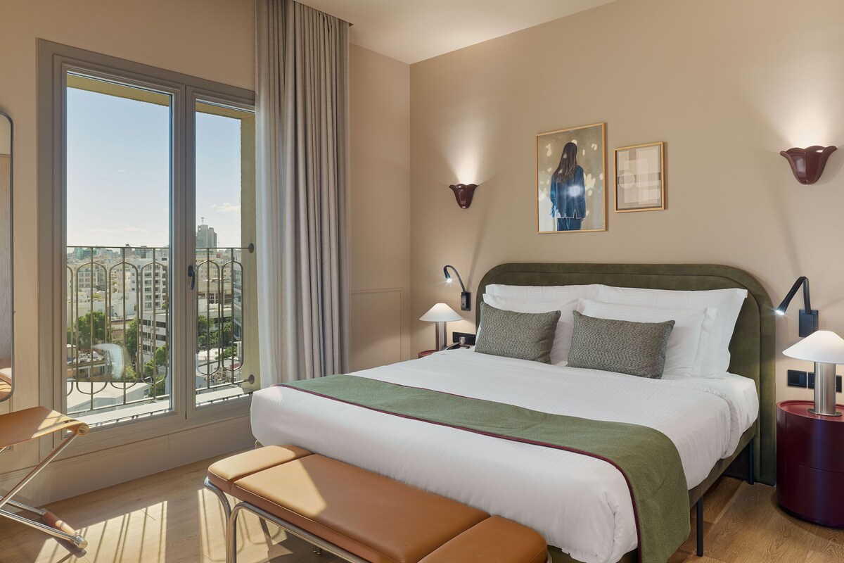 A hotel room with a large bed, a bench, a view of the city, and a cityscape visible through the window.