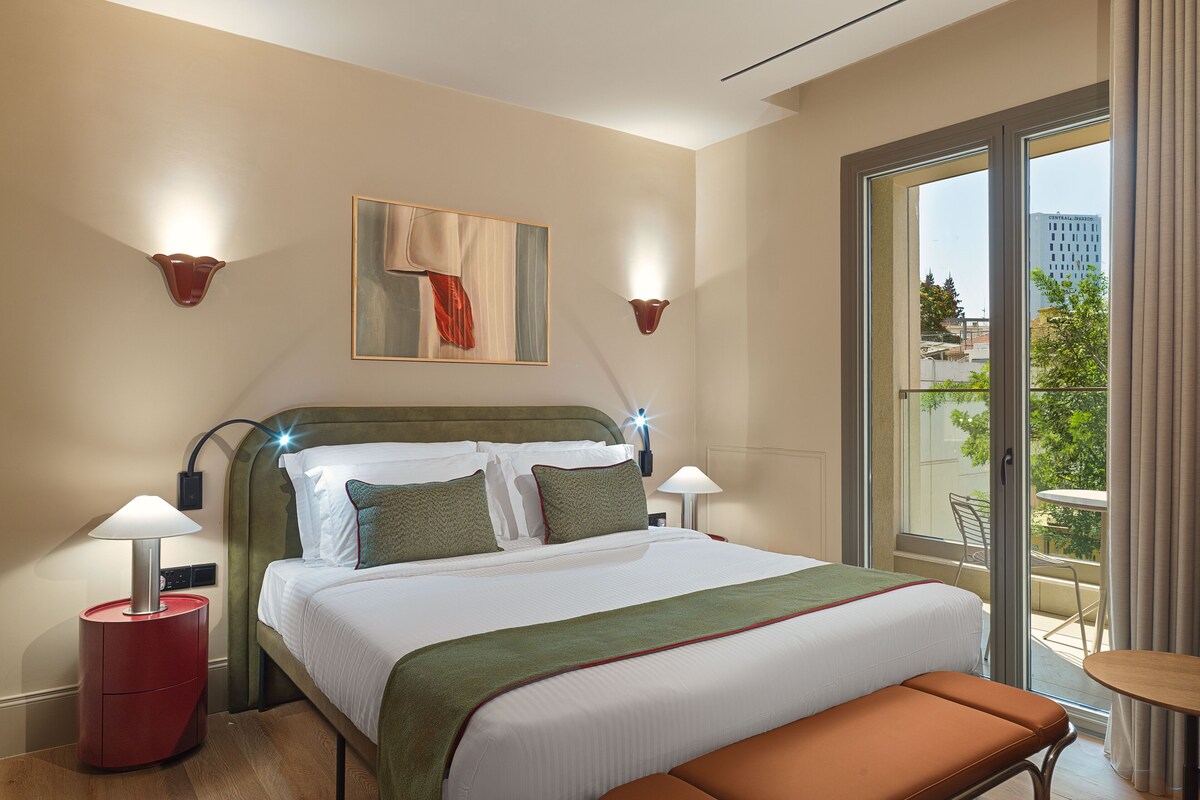 A hotel room with a bed, bedside lamps, a bench, a painting, and a view of the city.