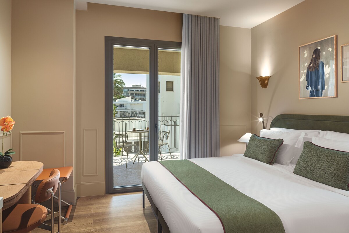 A hotel room with a bed, a balcony with chairs, a table, and a view of buildings outside.