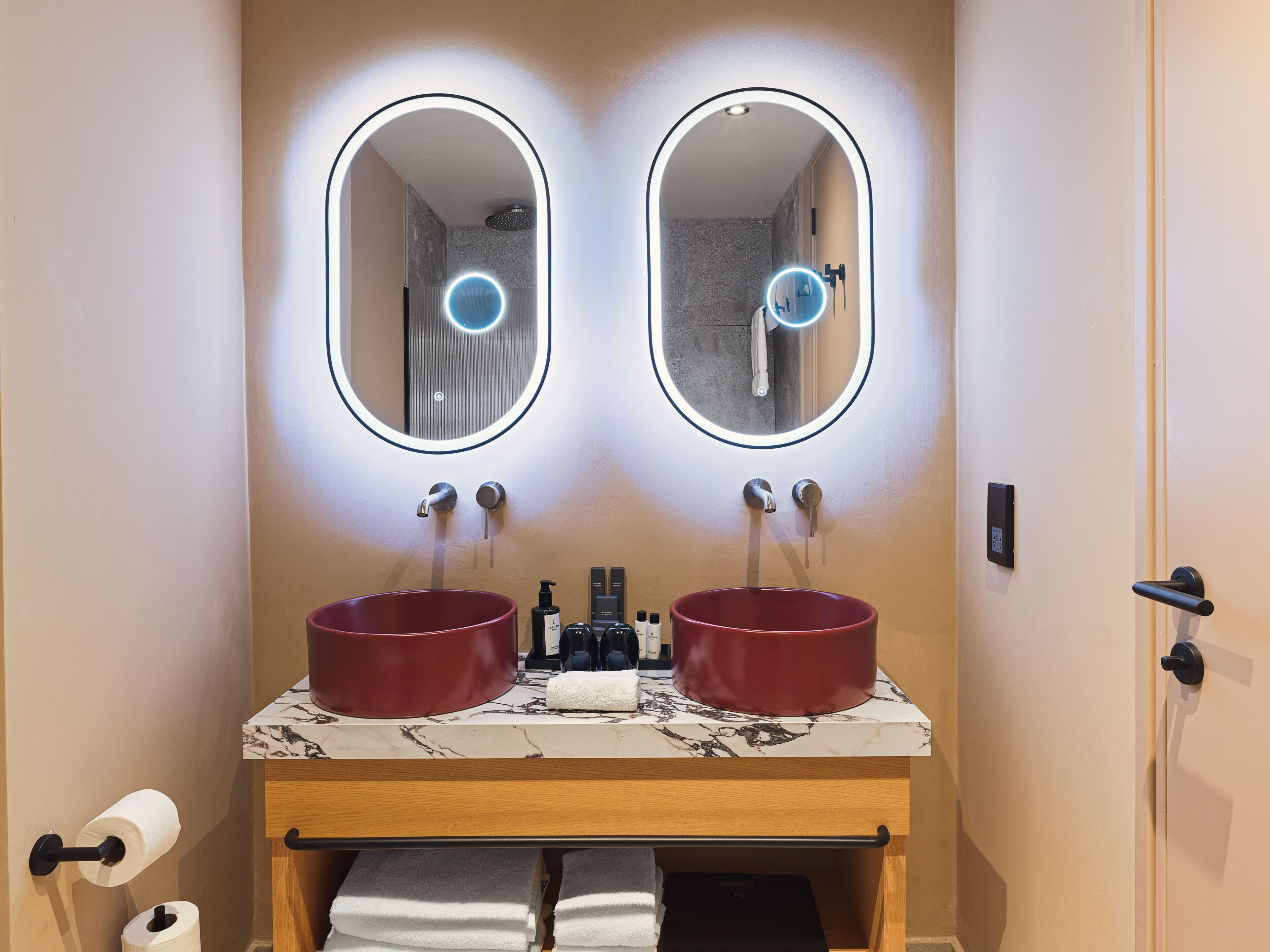 A bathroom with two sinks, each with a round mirror above. There is a towel rack below the sinks, and a toilet paper holder to the left.