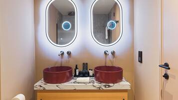 A bathroom with two sinks, each with a round mirror above. There is a towel rack below the sinks, and a toilet paper holder to the left.