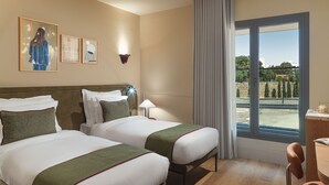 Quarto de hotel com duas camas, uma escrivaninha, uma cadeira, janela com vista e obras de arte na parede.
