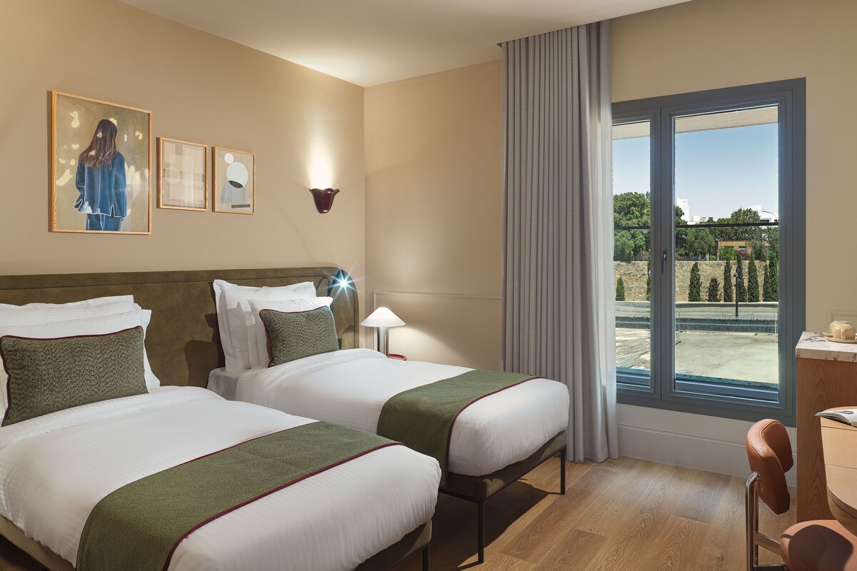 A hotel room with two beds, a desk, a chair, a window with a view, and artwork on the wall.