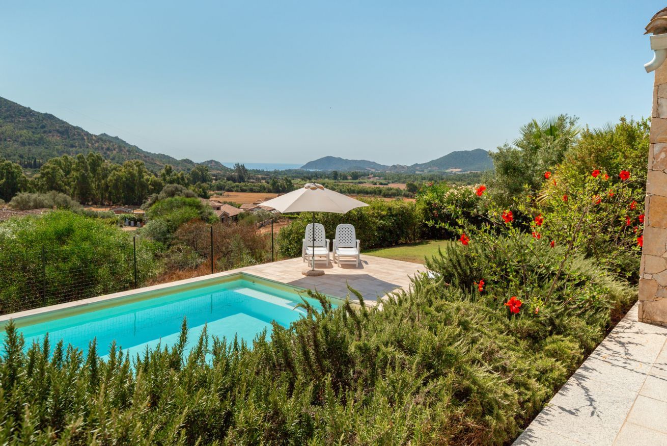 A swimming pool with two chairs and an umbrella, surrounded by greenery and a view of hills.