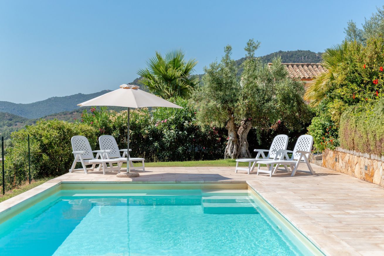A swimming pool with lounge chairs and an umbrella, surrounded by greenery and hills.