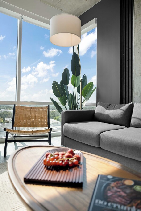 Ein modernes Wohnzimmer mit großem Fenster, einem grauen Sofa, einem Korbstuhl, einem Tisch mit Obstplatte und einer hängenden Pflanze.