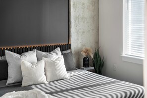 Une chambre à coucher avec un lit, des oreillers, une table de chevet, un vase contenant des plantes séchées et une fenêtre avec des stores.