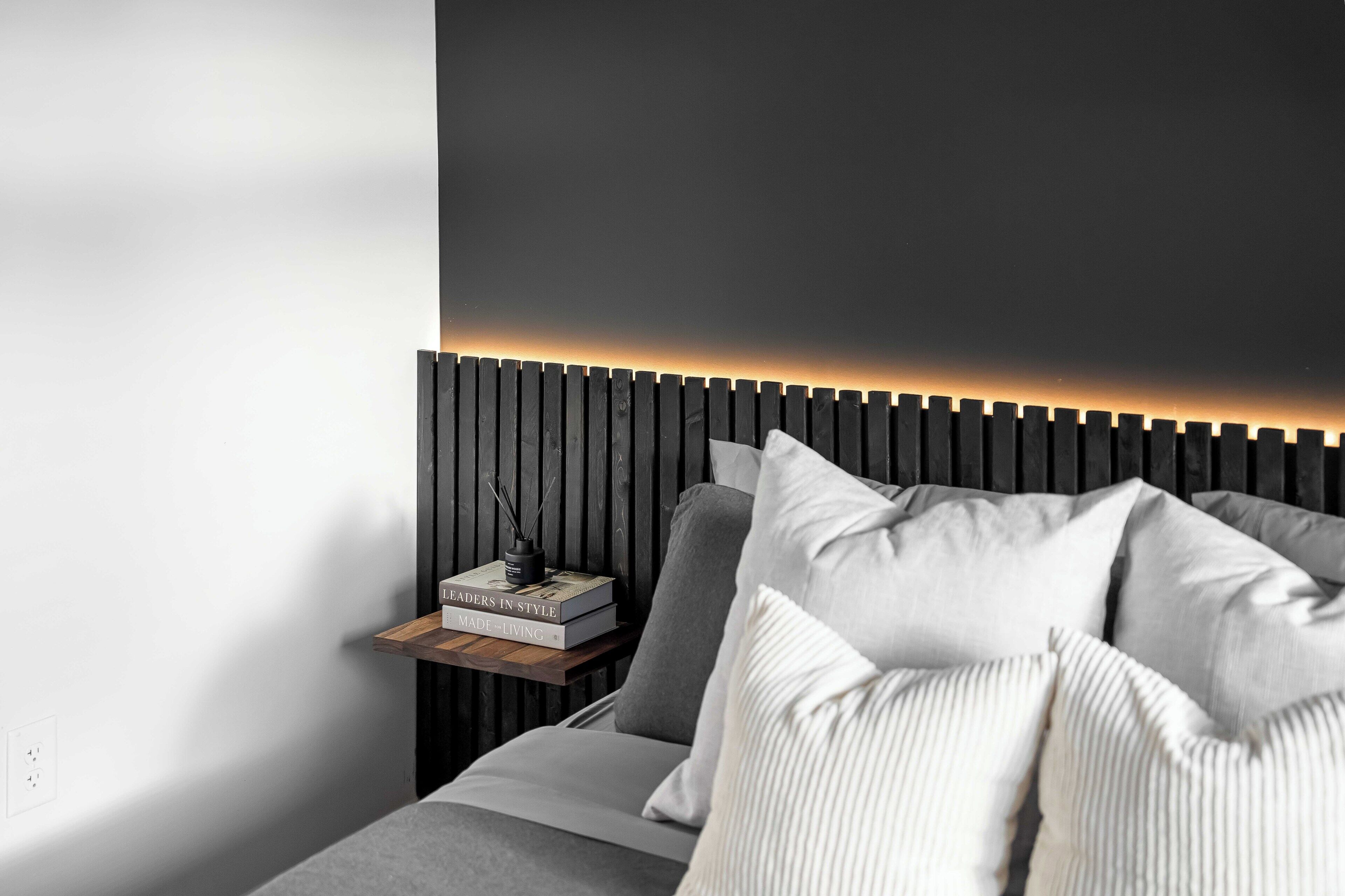 Un dormitorio moderno con cabecera oscura, mesita de noche con libros y lámpara de noche.