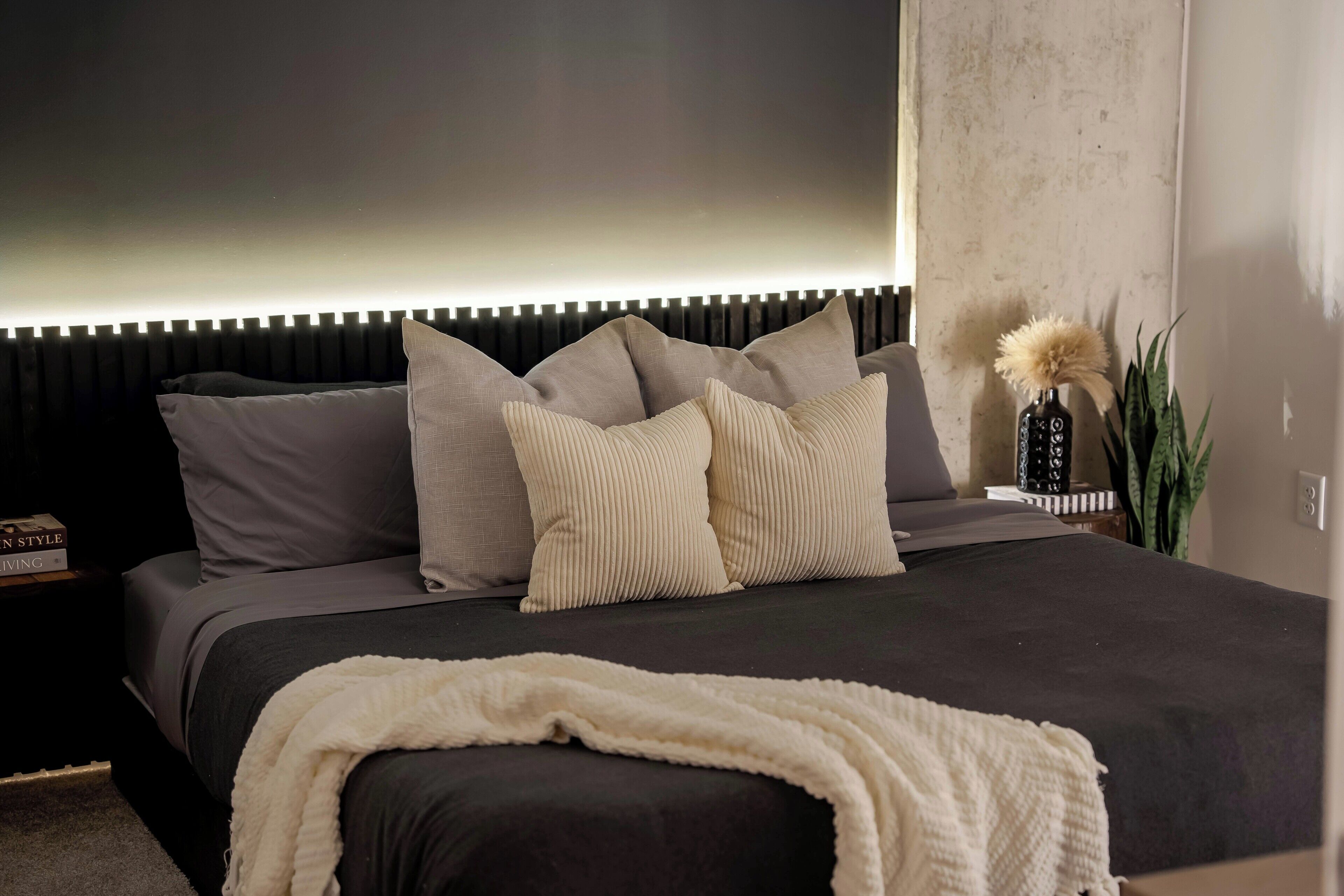 Een moderne slaapkamer met een donker bed, meerdere kussens, een nachtkastje met een boek en een decoratieve vaas met een plant.
